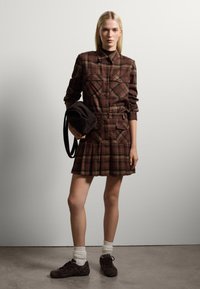 Braunes kariertes Kleid mit langen Ärmeln, zwei Brusttaschen und einem gefältelten Rock. Die Model hält eine braune Tasche und trägt weiße Socken mit dunklen Schuhen.
