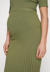 Kobieta w ciąży w zielonej, prążkowanej sukience z krótkimi rękawami, z rękami opartymi na brzuchu i biodrach.