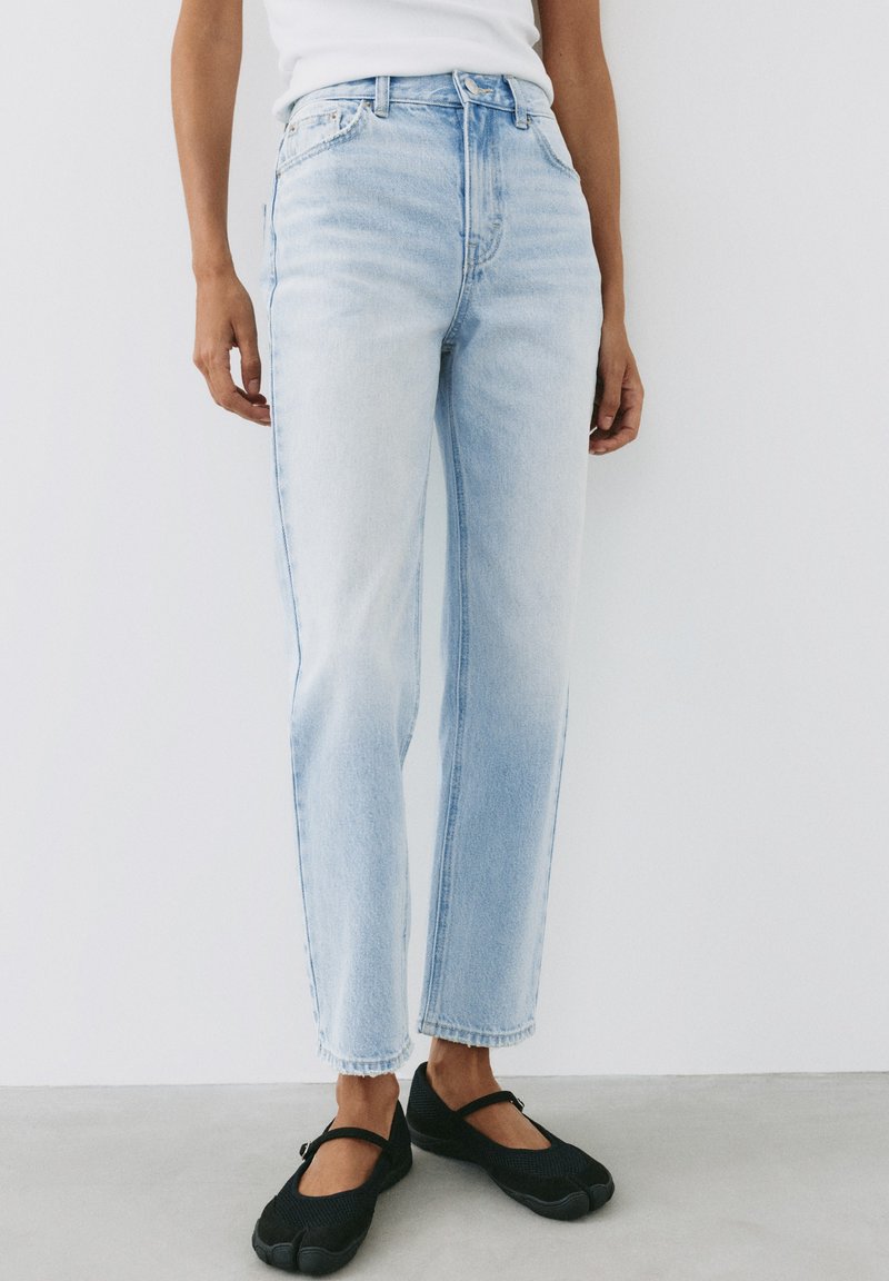 Personne portant un jean droit taille haute bleu clair associé à des chaussures style Mary Jane noires, debout devant un fond blanc.