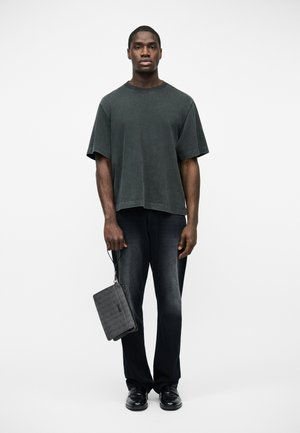 Young man in loose dark gray T-shirt, faded black jeans, black shoes, holding a patterned black wristlet bag against a plain white background.