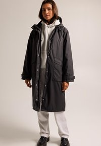 Parka imperméable noire avec capuche, dotée de boutons-pression, de deux poches avant et d'un cordon de serrage à la taille, superposée à un sweat à capuche gris.