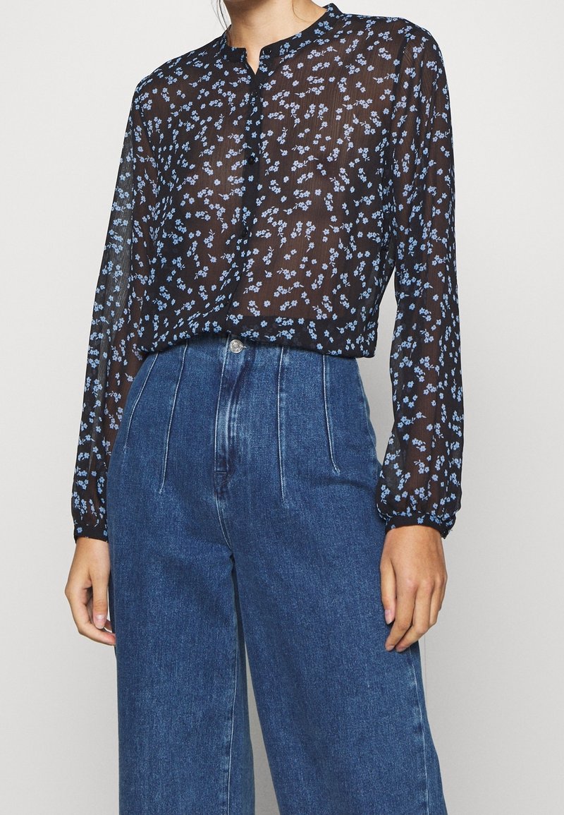Durchscheinende schwarze Bluse mit blauem Blumenmuster, langen Ärmeln mit gerafften Manschetten und einem geknöpften Dekolleté, kombiniert mit hochgeschnittenen Jeanshosen.