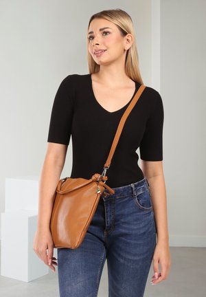 Junge Frau trägt ein schwarzes Kurzarmshirt und blaue Jeans, trägt eine braune Leder-Umhängetasche und steht vor einem schlichten Hintergrund.