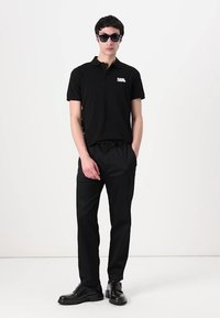 Čierny polokošeľa s logom, čierne nohavice s elastickým pásom a čierne slnečné okuliare. Model stojí na jednofarebnom pozadí.