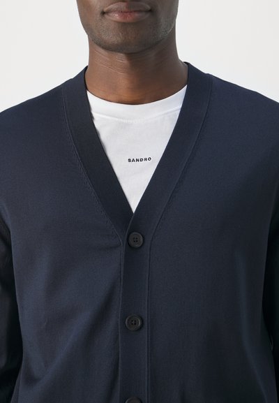 Cárdigan azul marino hecho de tejido suave con escote en V y cinco botones. Lleva puesto un T-shirt blanco que muestra la marca "SANDRO."