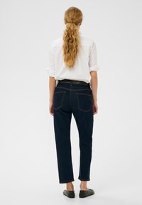 Frau mit langen, welligen Haaren, die zu einem Pferdeschwanz gebunden sind, trägt ein weißes Hemd, dunkelblaue Jeans mit Taschen, einen schwarzen Gürtel und schwarze Ballerinas. Sie steht mit dem Rücken zur Kamera.