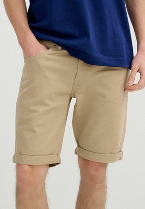 Hombre con pantalones cortos beige remangados y camiseta azul marino, con una mano en el bolsillo y la otra relajada a un lado.