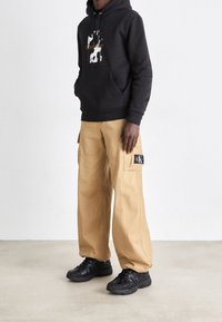 Uomo con felpa con cappuccio nera Calvin Klein, pantaloni cargo beige a gamba larga con patch CK e scarpe da ginnastica nere che sta in piedi su un pavimento chiaro contro uno sfondo bianco.