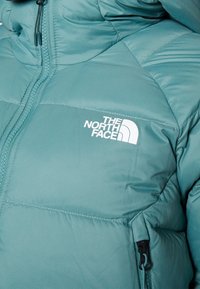 Veste matelassée bleu sarcelle avec le logo "The North Face" sur la poitrine et une fermeture éclair partiellement visible sur le côté gauche.