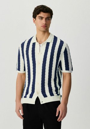 Mann trägt ein kurzärmeliges, geknöpftes Strickhemd mit vertikalen navyblauen und weißen Streifen, steht vor einem einfarbigen Hintergrund.