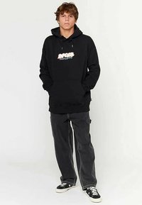 Black hoodie with a front pouch pocket, featuring a multi-colored graphic logo. Paired with dark grey baggy jeans and black sneakers.