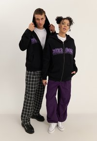 Deux jeunes adultes portant des sweat-shirts à capuche noirs avec des lettres violettes ; l'un porte un pantalon à carreaux et des chaussures noires, l'autre un pantalon violet et des baskets blanches.