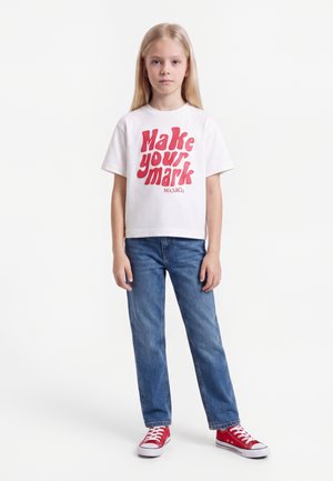 Młoda dziewczyna z długimi blond włosami, ubrana w białą koszulkę z czerwonym napisem "Make your mark", niebieskie dżinsy i czerwone trampki, stoi na białym tle.