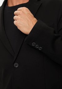 Blazer nero con revers a punta, tessuto testurizzato, tre bottoni neri sul polsino e una ripresa ravvicinata di una mano che tiene il rever.