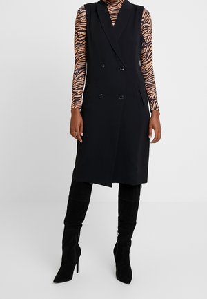 Femme portant un manteau noir sans manches à double boutonnage par-dessus un haut à manches longues imprimé tigre, associé à des bottes noires montantes à talons.