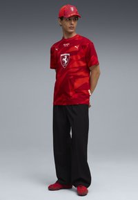 Rode T-shirt met geometrisch patroon en Ferrari-logo, gecombineerd met zwarte broek en rode sneakers, aangevuld met een bijpassende rode pet.