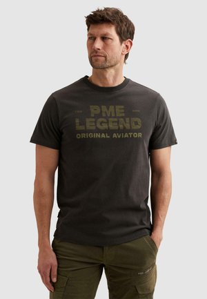 Άνδρας που φοράει μαύρο μπλουζάκι PME Legend με το κείμενο «Original Aviator» και ελιά cargo παντελόνι, στέκεται με τα χέρια εν μέρει στις τσέπες.
