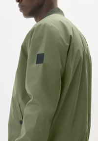 Olive green bomber jacket with a smooth texture, ribbed collar, and a logo patch on the left shoulder, featuring side pockets.