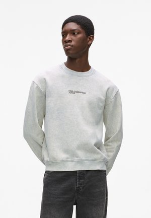 Szara, teksturowana bluza z okrągłym dekoltem i wyszywanym logo "KARL LAGERFELD JEANS" na piersi; połączona z ciemnymi dżinsami.