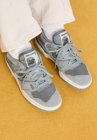 Nærbilde av føtter iført grå New Balance 550 sneakers med hvite sokker og beige bukser på et sennepsgult gulv.