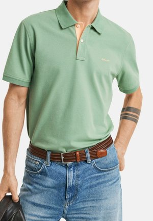 Polo vert avec un col orange contrastant, en coton texturé. Porté avec un jean en denim bleu et une ceinture tressée marron.