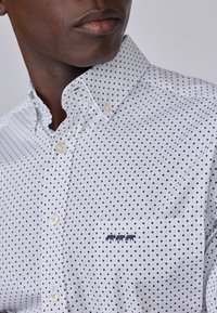 Chemise blanche à boutons avec un motif en diamants marine, comportant un détail de poche avec un design brodé bleu. Tissu en coton au toucher lisse.