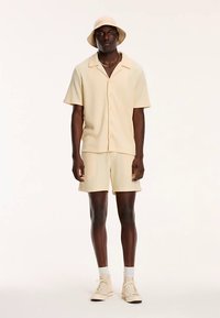 Camisa beige de manga corta con textura acanalada, combinada con pantalones cortos a juego y un sombrero tipo cubo, que presenta un ajuste relajado y una textura de tela suave.