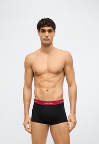 Juodi medvilniniai bokseriai su raudonais Calvin Klein logotipo juosmenimis, pasižymintys prigludusiu dizainu ir sklandžia tekstūra. Modelis stovi veidu į priekį.