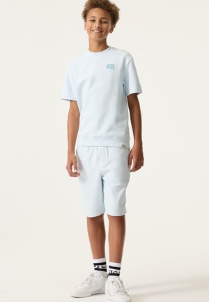 Hellblaues Baumwoll-T-Shirt mit kurzen Ärmeln und bedrucktem Text, kombiniert mit passenden Shorts. Weiße Sneakers mit schwarzen Details vervollständigen das Outfit.