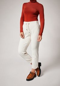 Pantalon blanc texturé avec taille élastique et cordon de serrage, associé à un col roulé rouge. Chaussures marron avec semelles noires complètent la tenue.