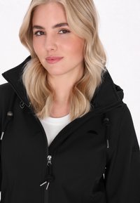 Schwarze Zip-Jacke mit hohem Kragen, ausgestattet mit Kordelzug und einem gestickten Regenschirm-Logo auf der Brust, getragen über einem weißen Hemd.