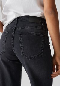 Black denim jeans featuring a fitted cut, back pockets with stitched detailing, and a plain black waistband. Smooth texture, no visible embellishments.