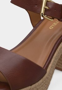 Sandale compensée en cuir marron avec boucle dorée et semelle en jute tressée, dotée d’une semelle intérieure lisse couleur camel et d’un design à bout ouvert.