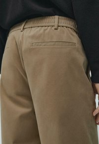 Kaki kalhoty mají elastický pas, boční kapsy a zadní kapsu, vyznačují se hladkou, strukturovanou látkou.