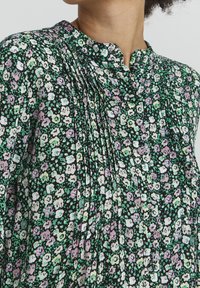 Blusa nera con motivo floreale, caratterizzata da un colletto alla mandarino, una texture plissettata e fiori verdi, viola e bianchi sparsi su uno sfondo scuro.