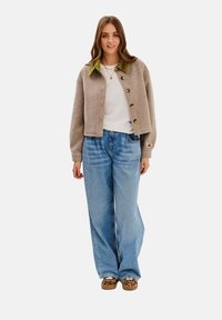 Brązowa krótka kurtka z zielonym kołnierzem i guzikami, noszona na białej koszuli, w połączeniu z szerokimi niebieskimi dżinsami i wzorzystymi brązowymi butami.