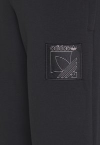 Černé tepláky mají texturovanou látku, s obdélníkovou černou nášivkou, na které je bílé logo Adidas a design tří pruhů.