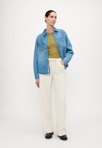 Blue denim jacket over an olive green patterned top, paired with cream wide-leg pants and brown shoes, against a plain white background.