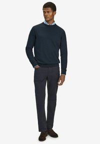 Marineblå stickad tröja med rundad halsringning, kombinerad med mörkgrå byxor. Outfiten har en ljusblå skjorta med krage under.