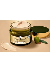 Un pot en verre vert avec un couvercle doré, contenant une crème beige clair. L'étiquette comprend des détails sur le produit et une branche d'olivier à côté du pot.