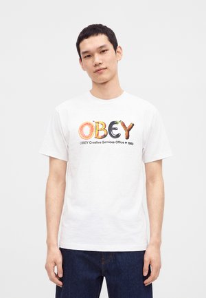 Mladý muž nosiace biele tričko s nápisom "OBEY" vytvoreným z písmen z ovocia a zeleniny a tmavomodré rifle na jednofarebnom pozadí.