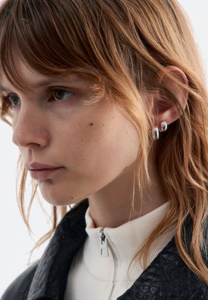 Silberne Ohrstecker an einem Modell mit schulterlangen, welligen braunen Haaren. Sie trägt ein hochgeschlossenes weißes Oberteil und eine strukturierte schwarze Jacke.
