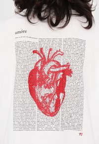 Hvit t-skjorte med rød hjerteillustrasjon og tekst på italiensk, med en blanding av kursiv og serif-skrifttyper som skaper en collage-effekt.