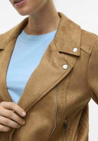 Veste en daim beige avec un col à revers, boutons-pression argentés et fermeture éclair latérale. Portée sur un t-shirt bleu clair. Texture lisse.
