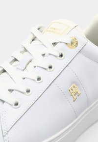 Sapatilha de couro branca com emblema dourado, atacadores brancos e ilhós redondos. Textura suave com design minimalista e detalhes de marca.