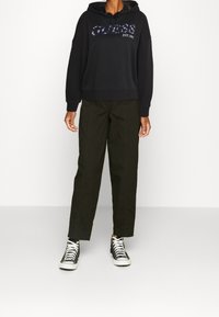 Sudadera negra con capucha y logo impreso, combinada con pantalones rectos de color verde oscuro y zapatillas de deporte negras de caña alta. Costuras de contraste visibles.