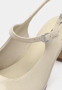 Krémpová lakovaná kůže Mary Jane bota se stříbrnou sponou a nízkým blokovým podpatkem na bílém pozadí.