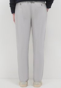 Persona che indossa pantaloni dritti grigio chiaro e scarpe beige, vista da dietro su uno sfondo bianco neutro.