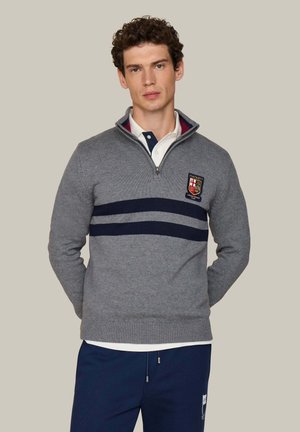 Hombre con un suéter gris con cremallera hasta la mitad, dos rayas azul marino y un escudo, camisa blanca debajo y pantalones azul marino sobre un fondo liso.