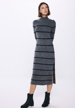 Graues Strickkleid mit schwarzen horizontalen Streifen, hohem Kragen, langen Ärmeln, seitlichem Schlitz und Knopfdetail an der Schulter, kombiniert mit schwarzen Knöchelstiefeln.